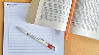 Prüfungssituation mit Schreibpapier, Stiften und einem aufgeschlagenen Buch. (verweist auf: Alles rund um das Thema Prüfungen)